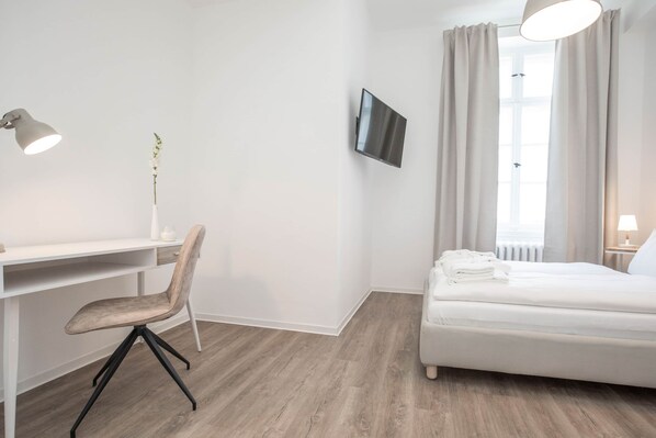 Ein Hotelzimmer mit einem Bett, einem Schreibtisch mit Stuhl, einem an der Wand montierten Fernseher und einem Fenster mit Vorhängen.