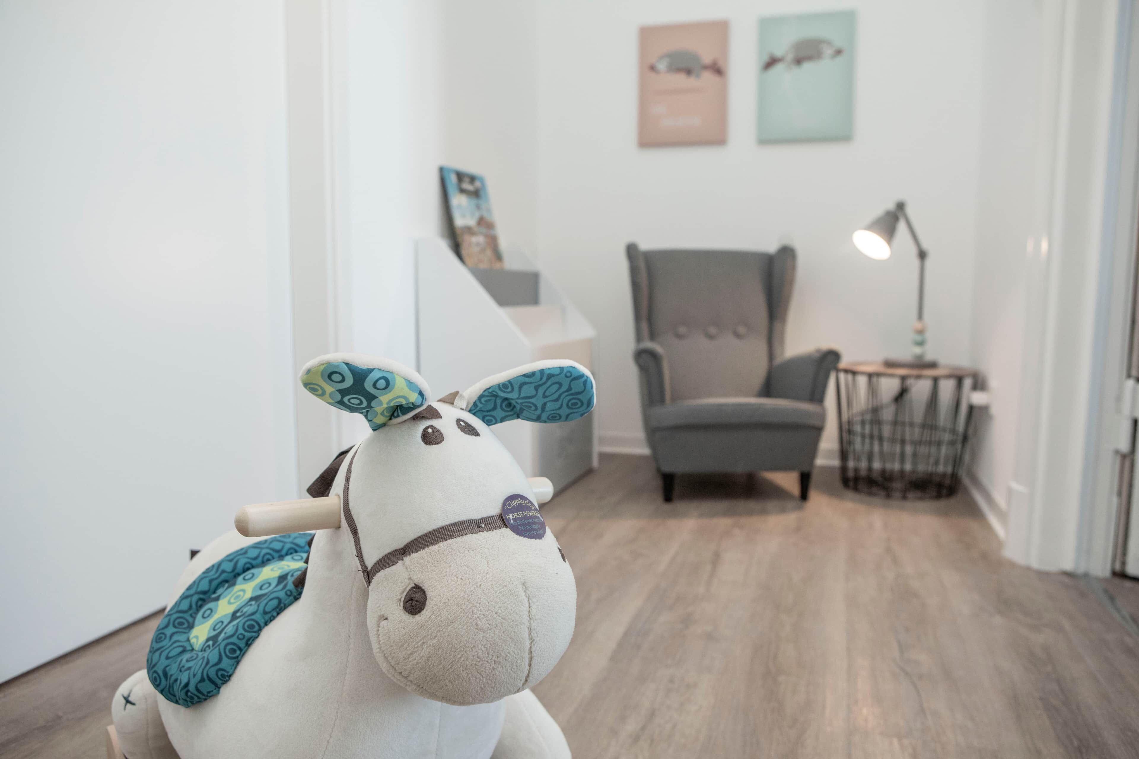A plush toy in a room with a grey armchair, a small table with a lamp, and a staircase in the background.
