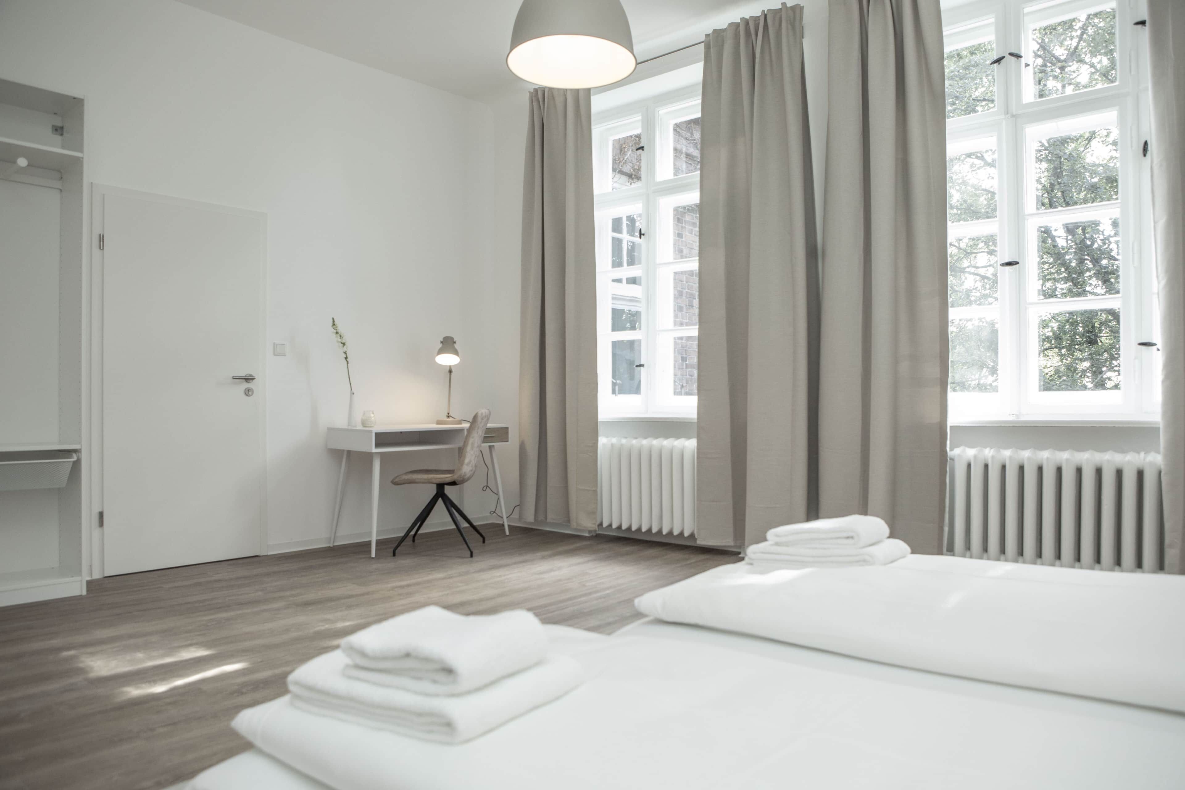 A modern hotel room with a bed, a desk, and a chair. There is a window with curtains and a view of trees outside.