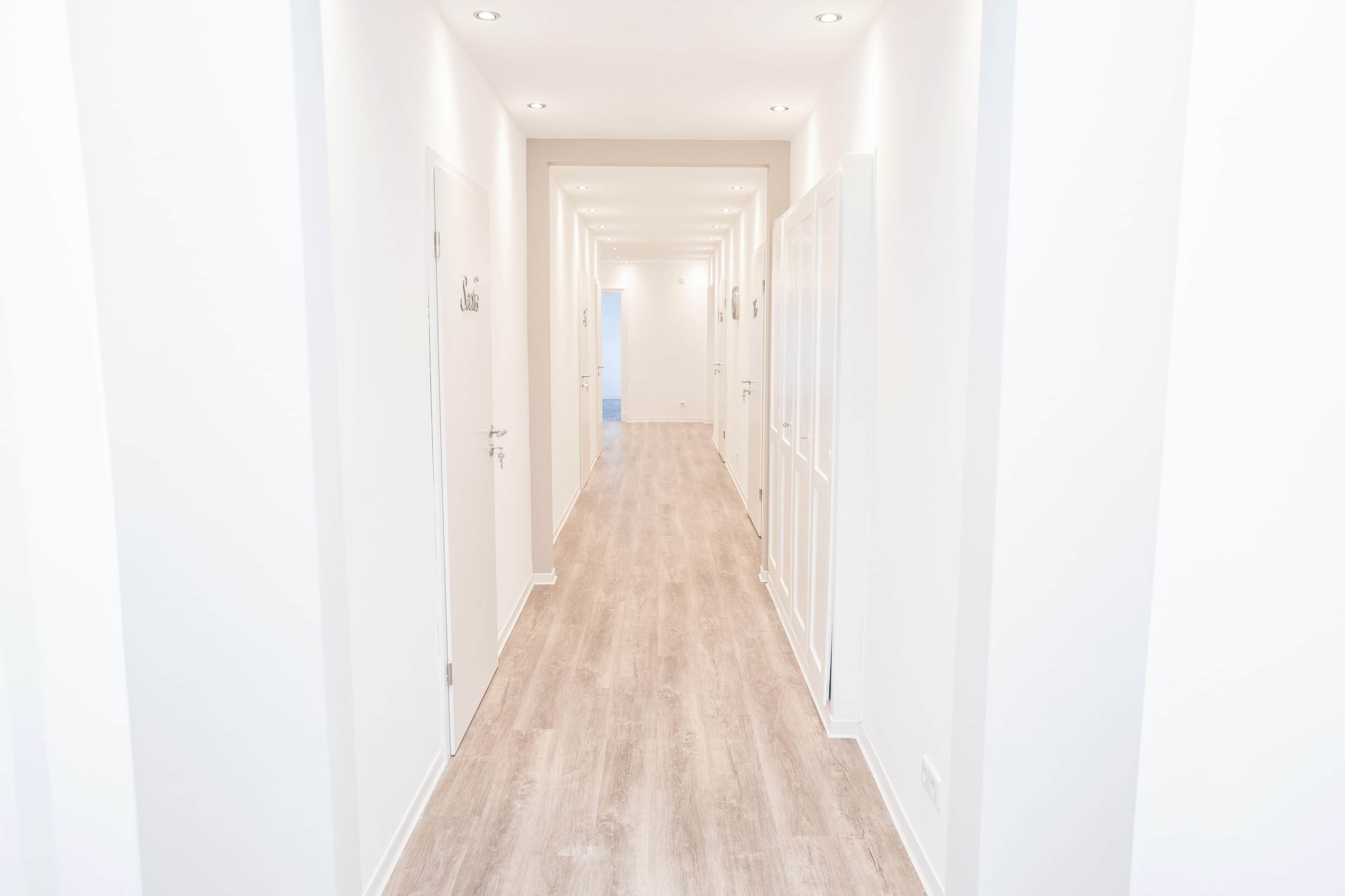 A long, narrow hallway with white walls and wooden flooring, leading to a door at the end.
