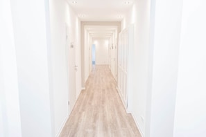 Un long couloir étroit aux murs blancs et au plancher en bois, qui mène à une porte au bout.