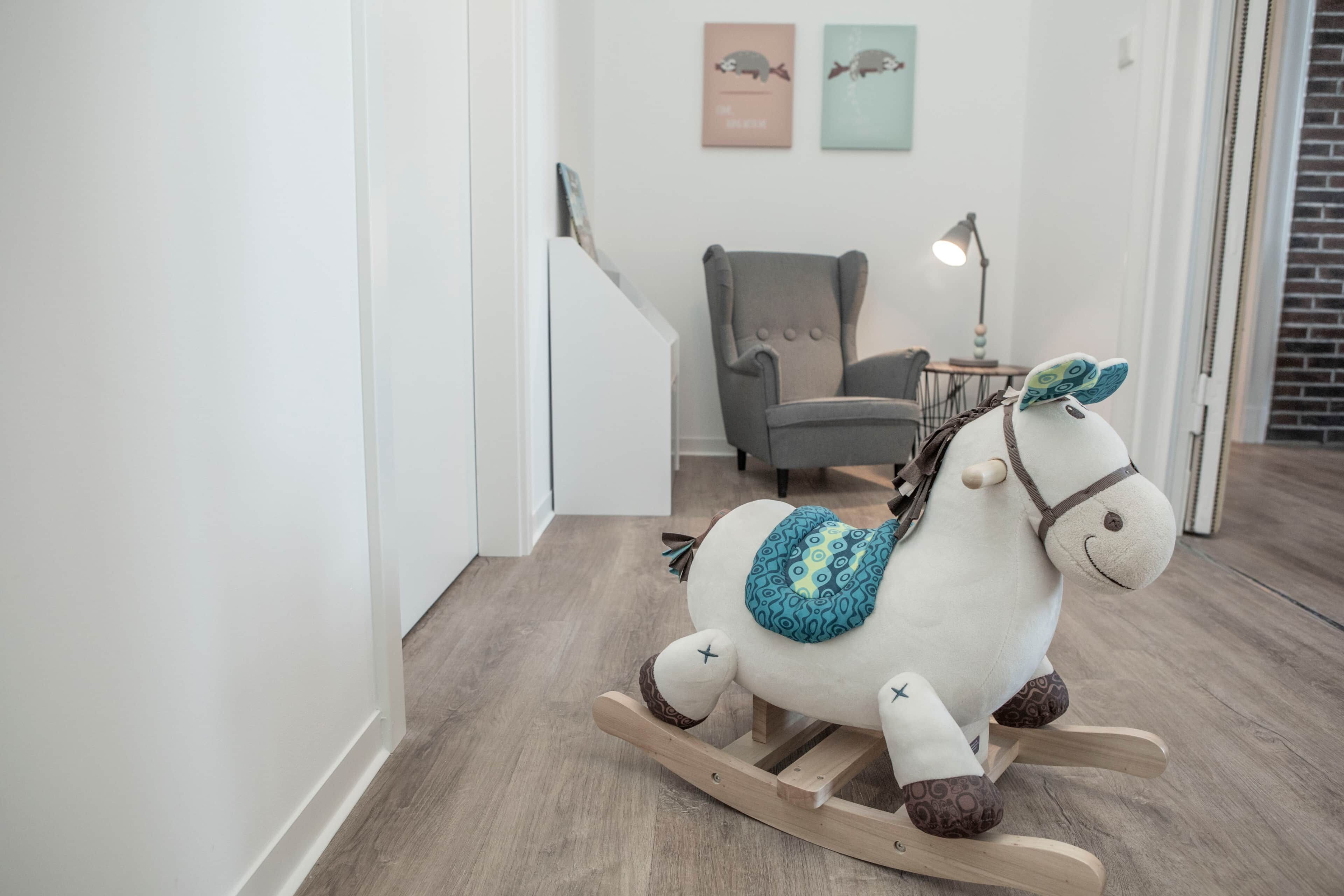 Ein hölzernes Schaukelpferd mit Sattel und dekorativen Elementen in einem Raum mit einem grauen Sessel, einer Lampe und einer Treppe.