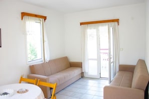 Ein Zimmer mit einem beigefarbenen Sofa, einem kleinen runden Tisch mit weißem Tuch und einem Fenster mit Vorhängen.