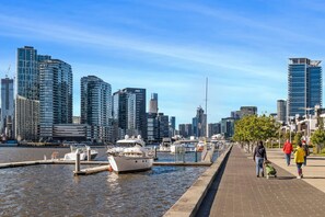 Un paisaje urbano frente al agua con modernos edificios altos, un puerto deportivo con barcos y un paseo peatonal.