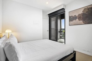 Un dormitorio con cama, mesita de noche, lámpara, puerta corrediza a un balcón y un cuadro en la pared.