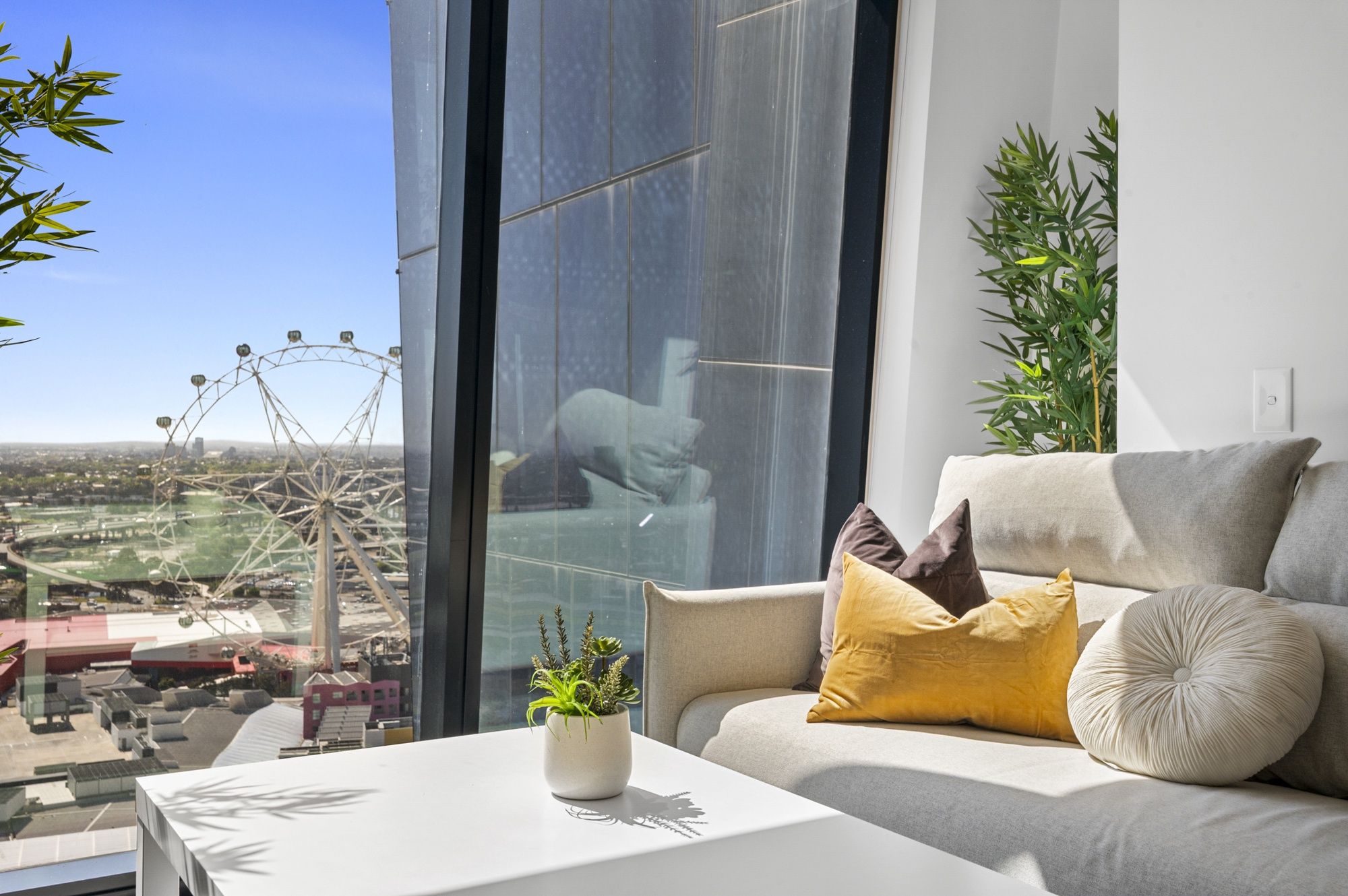 Een moderne woonkamer met een groot raam, een bank met kussens, een klein tafeltje met een plant en uitzicht op een reuzenrad.