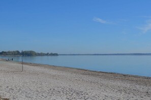 A beach with a clear sky, calm water, and a distant shoreline with trees and buildings. - Shell in House Fördeglück (Glücksburg)