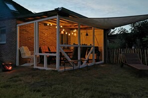 A covered outdoor seating area with a brick wall, white chairs, and hanging lights. - Holiday apartment with 3 bedrooms (Wittmund)