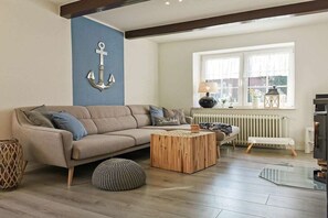 A living room with a beige sofa, a wooden coffee table, a patterned ottoman, a blue accent wall, and a large window with curtains. - Holiday apartment with 3 bedrooms (Wittmund)