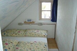Ein kleiner, schmaler Raum mit einem Bett, einem Fenster mit Vorhängen und einem Regal mit einem Bild und einem kleinen Gegenstand.