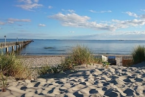 A sandy beach with a wooden pier extending into calm waters, a small structure with striped awnings, and sparse vegetation. - Holiday apartment with 1 bedroom (Lubmin)