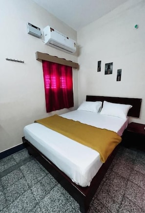 Habitació d'hotel amb un llit, una cortina vermella, un aparell d'aire condicionat i una petita prestatgeria penjada a la paret.