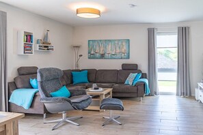 A modern living room with a sectional sofa, a grey armchair, a small ottoman, and a coffee table. There is a bookshelf with books and decorative items, a painting of sailboats on the wall, and a large window with curtains. - Holiday home Sailing view Luv (Glücksburg)