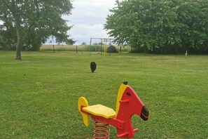 Ein grasbewachsenes Areal im Freien mit roter und gelber Spielausstattung, einer Schaukel und einem großen Baum im Hintergrund.