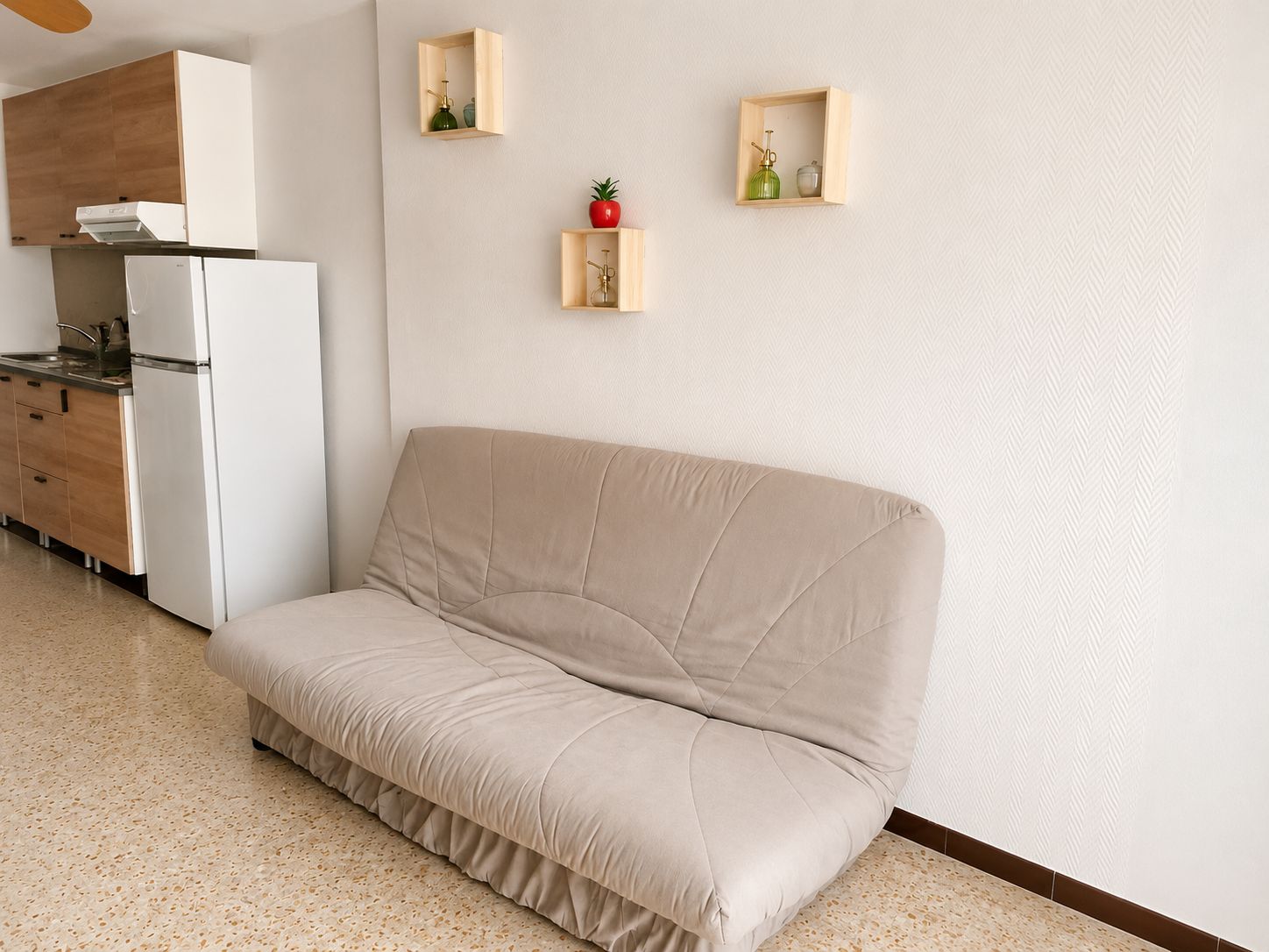 A modern living room with a beige sofa, a kitchen area with a white refrigerator, and two wooden shelves with decorative items.