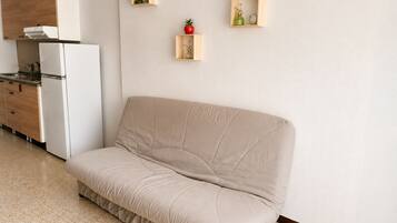 A modern living room with a beige sofa, a kitchen area with a white refrigerator, and two wooden shelves with decorative items.