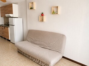 Un salon moderne avec un canapé beige, un coin cuisine doté d’un réfrigérateur blanc et deux étagères en bois ornées d’objets décoratifs.