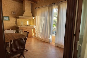 A kitchen with a wooden table and chairs, a brick backsplash, and a view through a curtain to an outdoor area. - Apartment with 1 bedroom (Chia SU, Domus de Maria)