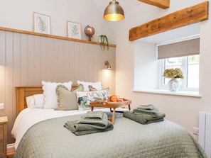 A neatly made bed with pillows and a blanket, a bedside table with a lamp, a window with a view, and a shelf with decorative items. - Tawny Cottage (Scarborough)