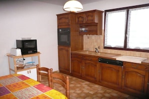 A compact kitchen with wooden cabinets, a microwave, an oven, a sink, and a window with lace curtains. - Lambezen-maison aux volets bleus (Camaret sur mer)