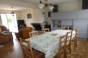 Een eetruimte met een tafel gedekt voor vier personen, omringd door houten stoelen. Achtergrond is een open haard zichtbaar, en links staat een televisie op een standaard.