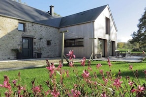 A stone building with a modern extension, a covered outdoor seating area, and a well-maintained lawn with flowering plants. - Holiday home with 1 bedroom (Les Moitiers-d'Allonne)