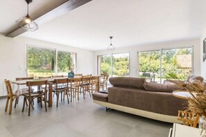 A spacious living area with a sofa, dining table, and chairs, large windows offering a view of greenery. - Holiday home with 1 bedroom (Les Moitiers-d'Allonne)