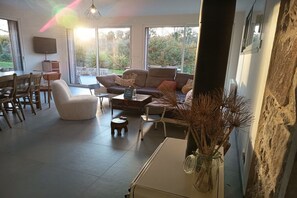 A spacious living area with a sofa, coffee table, and dining set. There is a large window providing a view of the outdoors. - Holiday home with 1 bedroom (Les Moitiers-d'Allonne)