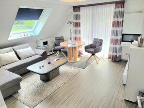A modern living room with a sofa, a coffee table, and two armchairs. There is a small table with a microwave and a lamp. The room has a large window with curtains and a view of greenery. - Apartment 'Nordseeperle Strandkorb Am Strand' with Balcony and Wi-Fi (Wangerland)