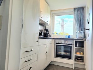 A compact kitchen with white cabinets, a built-in oven, and a window with a view of the sky. - Ferienwohnung 'Nordseeperle Strandkorb Am Strand' mit Balkon und WLAN (Wangerland)