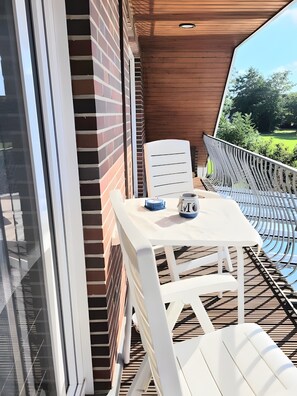 A balcony with a white table and chairs, a blue object on the table, and a view of greenery. - Ferienwohnung 'Nordseeperle Strandkorb Am Strand' mit Balkon und WLAN (Wangerland)