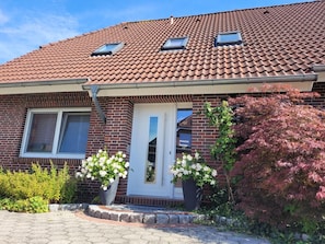 A residential building with a red-tiled roof, a brick facade, and a front door with a small window. There are potted plants with white flowers in front of the door. - Apartment 'Nordseeperle Strandkorb Am Strand' with Balcony and Wi-Fi (Wangerland)