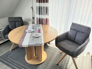 Pyöreä pöytä, jossa on puinen jalka, kaksi harmaata, verhoiltua tuolia, kuviollinen pöytäliina ja pari mukea pöydällä.