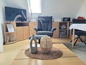 A living room with a blue armchair, a wooden cabinet, a television, and a small table with a red object on it. - Apartment 'Kleine Krabbe Strandkorb Am Strand' with Balcony and Wi-Fi (Wangerland)