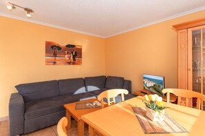 A living room with a sofa, a dining table with chairs, a television, and a cabinet. - Holiday apartment Haus Atlantik 319552 (Kühlungsborn - Ost)