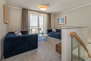 A modern living room with a dark blue sofa, a round coffee table, and a view of the outdoors through large windows. - Holiday apartment with 1 bedroom (Lubmin)