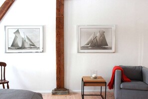 A room with a wooden beam, two framed pictures of sailboats, a small table with a candle, a grey sofa, and a wooden chair. - Sonnenhof by the sea (Osterhever)