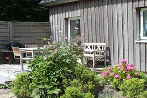 A wooden cabin with a deck, outdoor seating, and flowering plants. - Sonnenhof by the sea (Osterhever)