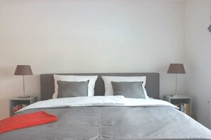 Un letto ben rifatto con un piumone grigio e cuscini, due comodini con lampade e una coperta rossa.