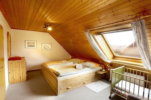 Une chambre avec un plafond en pente, un lit, un berceau, une fenêtre avec des rideaux et une armoire en bois.