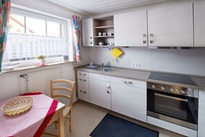 A compact kitchen with white cabinets, a built-in oven, and a window with a curtain. - FH Hus achter´n Diek (Hooksiel)