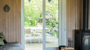 Habitación con cama, suelo de madera, puerta corrediza de vidrio que da a un área exterior con mesa y sillas, y estufa de leña.