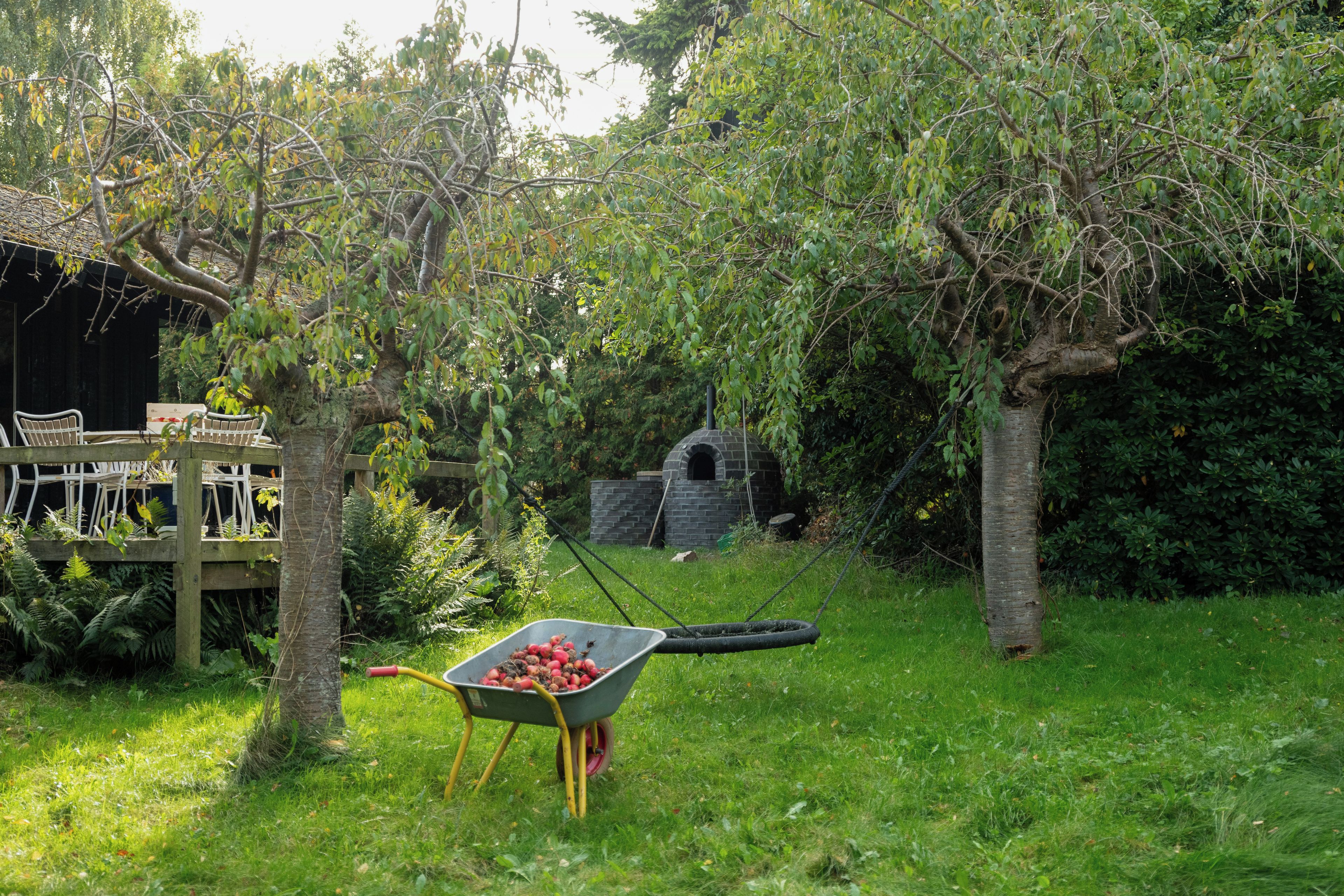 Un jardín con un carretón, una terraza de madera y una estructura de piedra.