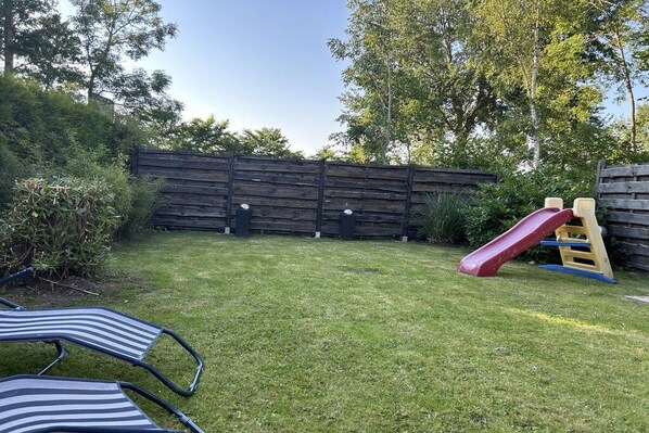 Ein Garten mit Holzzaun, Rutsche und zwei Streifenliegestühlen.