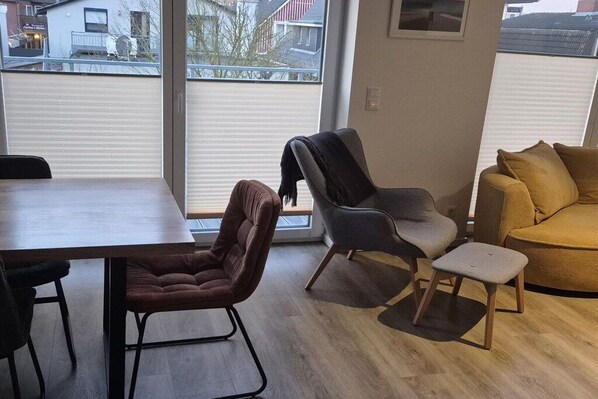 A modern living room with a dining table, two chairs, a sofa, and a window with a view of a residential area. - Holiday apartment in Büsum with swimming pool (Büsum)