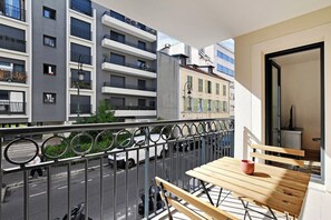 Ein Balkon mit Holztisch und -stühlen, Blick auf eine Straße mit Autos und ein Gebäude im Hintergrund mit mehreren Balkonen.
