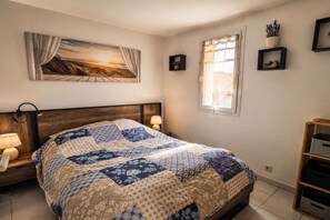 A bedroom with a wooden headboard, a quilted bedspread, bedside lamps, a window with curtains, and wall-mounted shelves with decorative items. - Holiday apartment with 1 bedroom (Leucate)