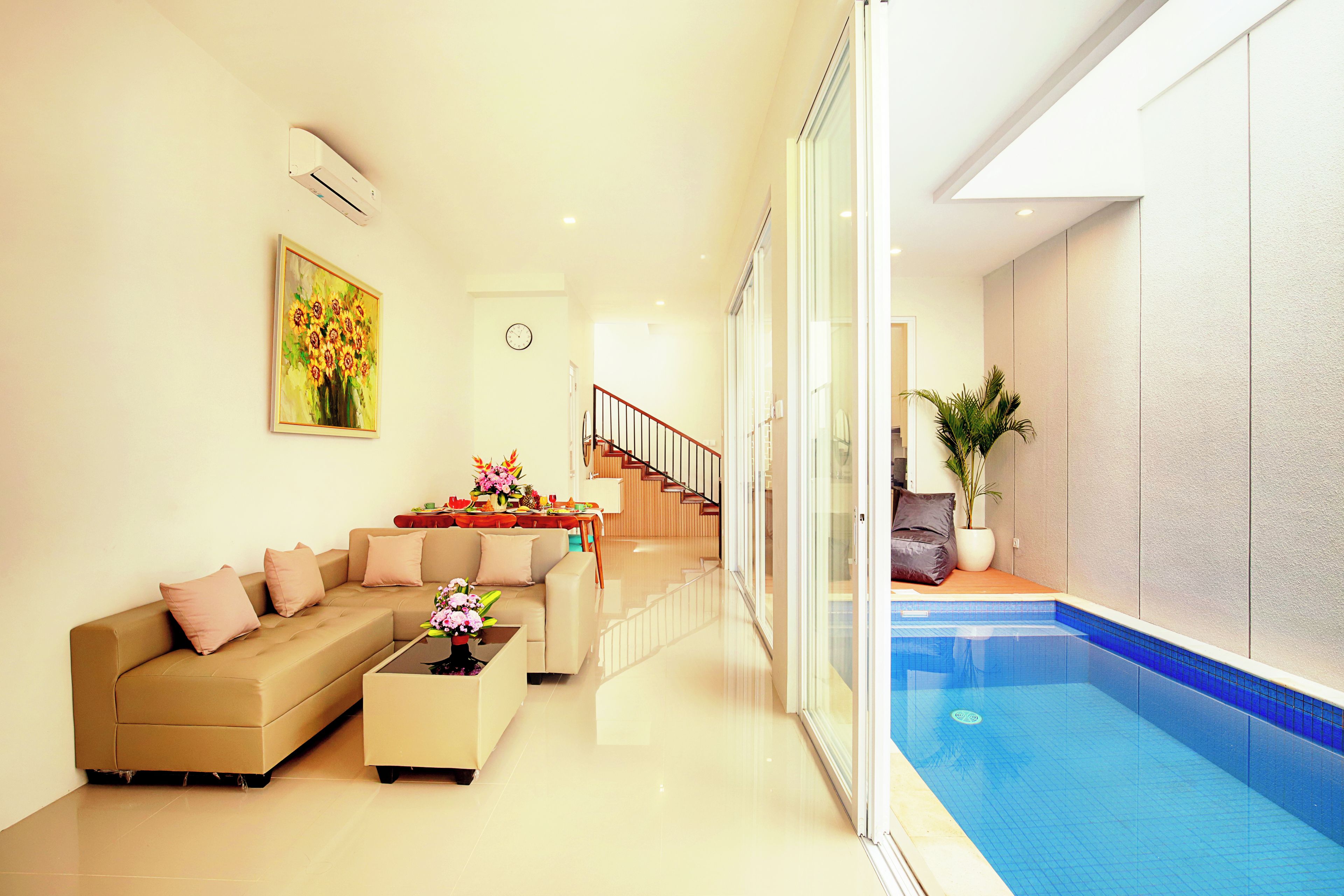 A modern living room with a sofa, coffee table, and a painting of sunflowers. There is a staircase leading to another level, and a glass door to a swimming pool.