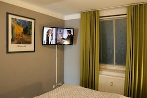 Ein Hotelzimmer mit einem Bett, einem an der Wand montierten Fernseher, einem gerahmten Bild und einem Fenster mit Vorhängen.