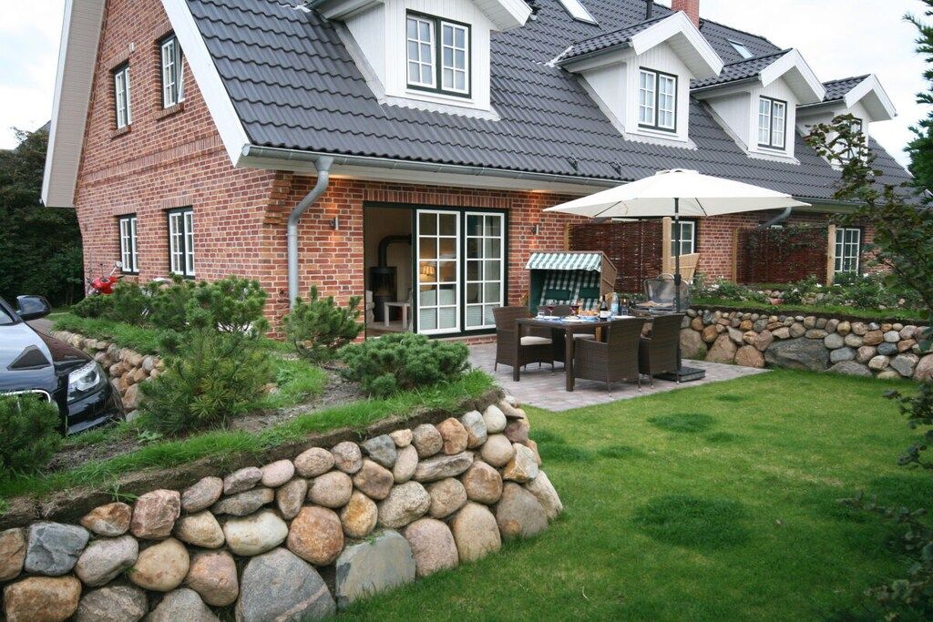 A two-story house with a stone retaining wall, a patio with outdoor furniture, and a car parked in the driveway.